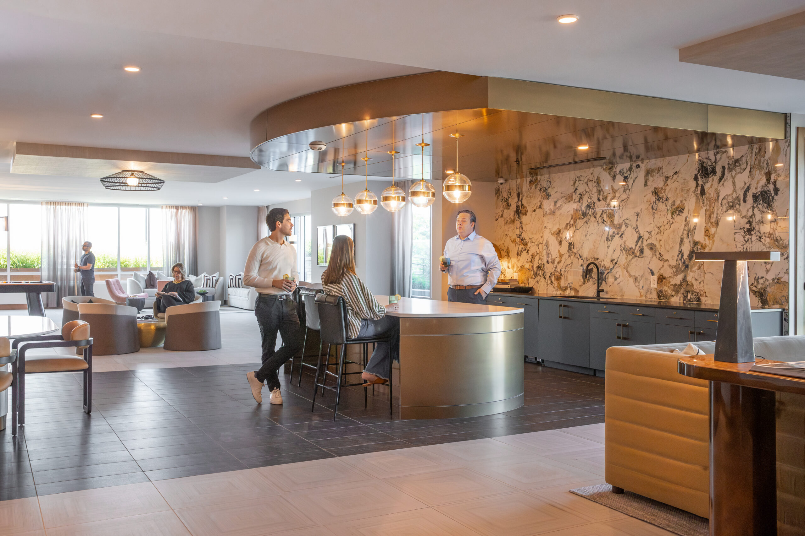 Woman sitting talking to a man in the kitchen area of the furnished and decorated clubroom. Woman in the background reading. Two men in the background plating pool.